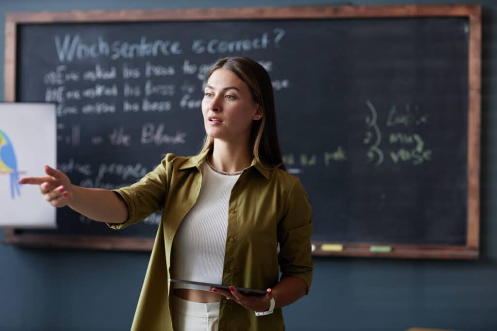 Teacher standing in front of a blackboard, holding a tablet, teaching a class